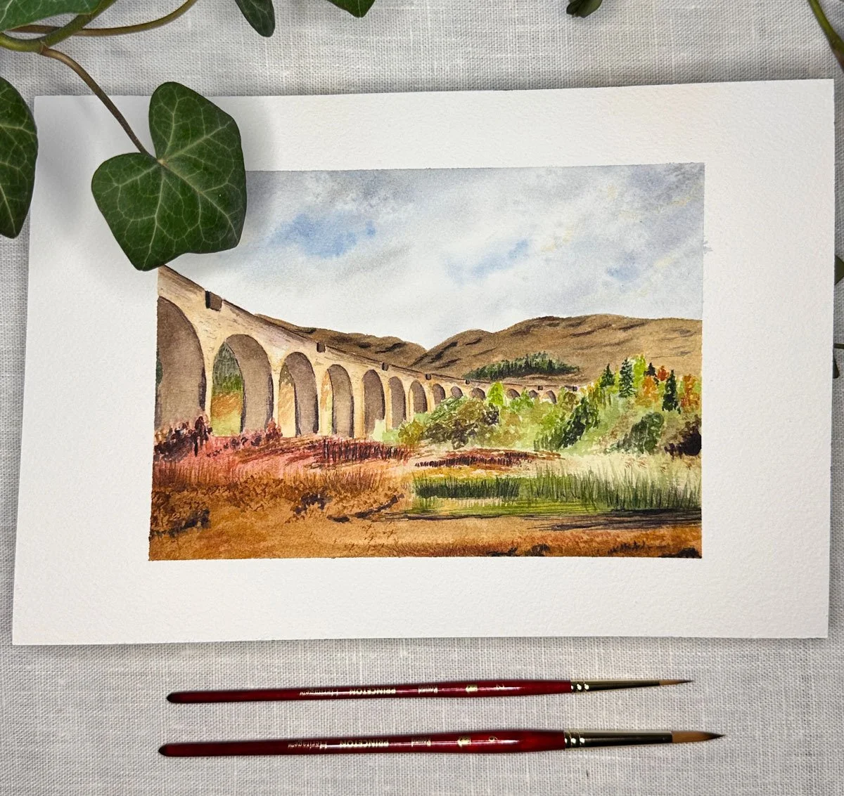 A watercolor painting of a landscape featuring a stone viaduct with multiple arches, rolling hills in the background, and lush trees and shrubs in the foreground. The painting is framed with a white border and placed on a white surface, accompanied b