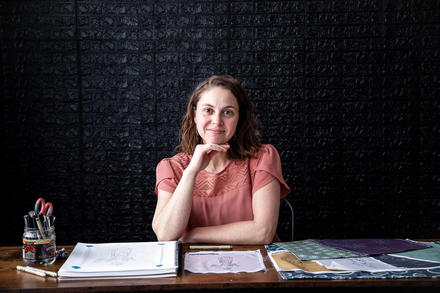 A woman with brown, shoulder-length curly hair sitting at a wooden table with fabric swatches, sketchbook, pens, and scissors, smiling at the camera against a black brick wall background.
