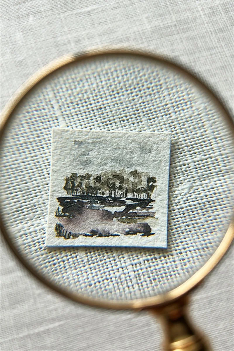 A small painting of trees and a river, viewed through a magnifying glass.