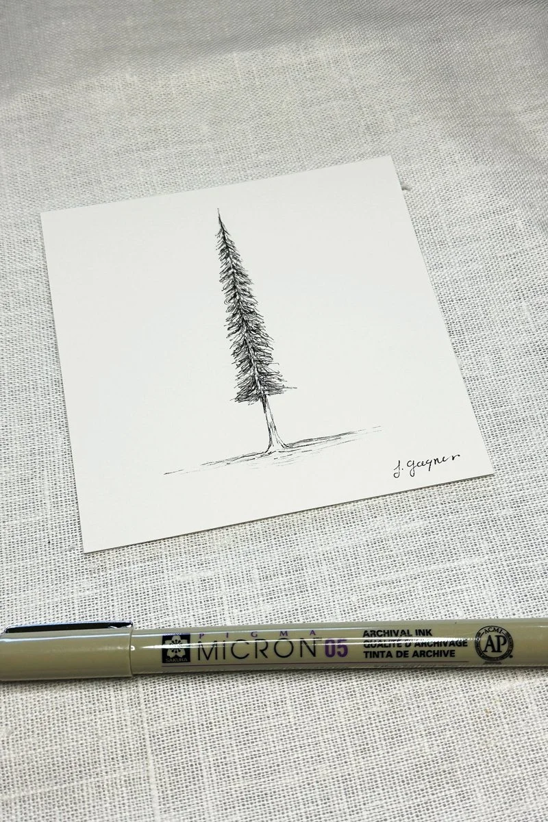 A simple black ink sketch of a tall, narrow pine tree on a white sheet of paper. The sketch is signed by J. Geynar, and a beige Micron pen lies below the paper on a textured surface.