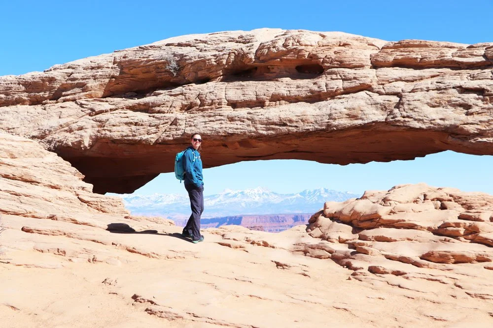 Person standing under a large, natural rock arch in a desert landscape with snow-capped mountains in the distance.