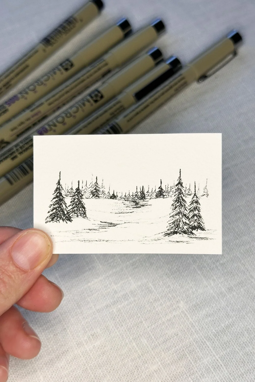 A hand holding a small sketch of a winter landscape with snow and pine trees, with several black pens in the background on a white surface.