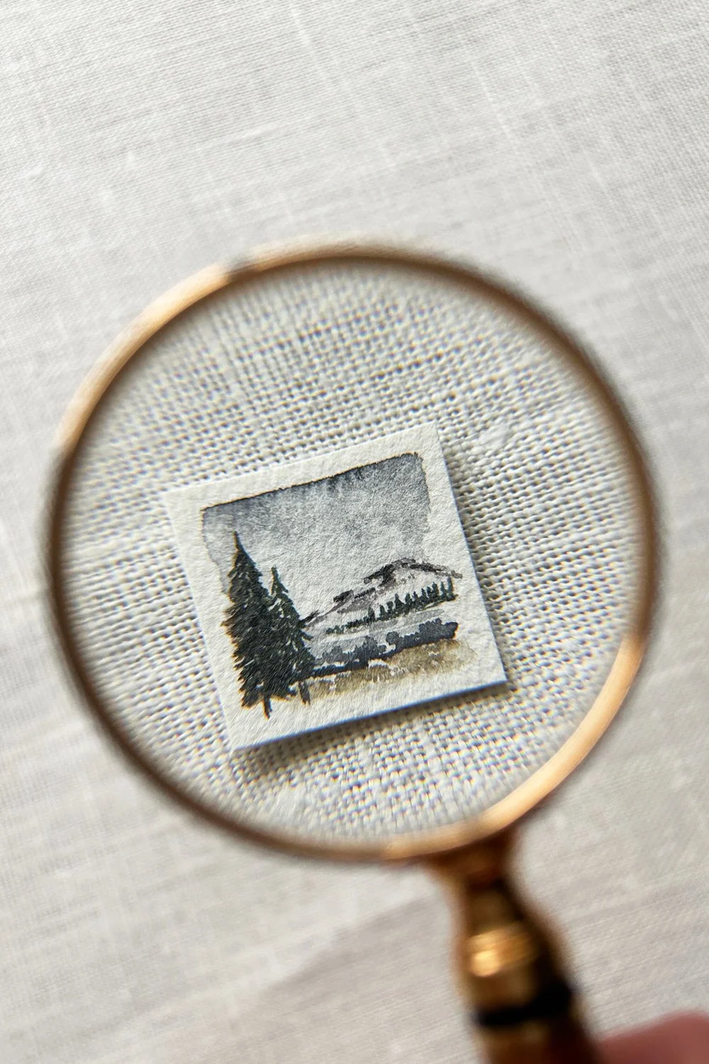 A small watercolor painting of a landscape with trees, hills, and a cloudy sky, viewed through a magnifying glass.