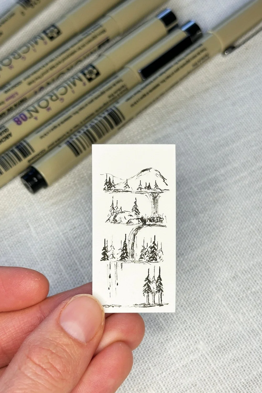 A hand holding a small sketch pad with a simple ink drawing of mountains and trees, with markers visible in the background.