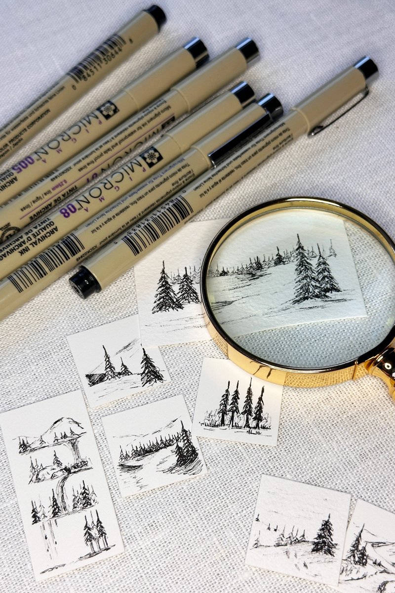 A collection of black ink sketches of landscapes with trees, a mountain, and water, some of which are being viewed through a magnifying glass, along with five Micron pens on a white textured surface.