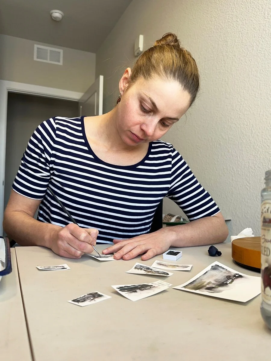 Woman with brown hair tied in a bun, wearing a navy and white striped shirt, is sitting at a table, carefully handling small black and white photographs.
