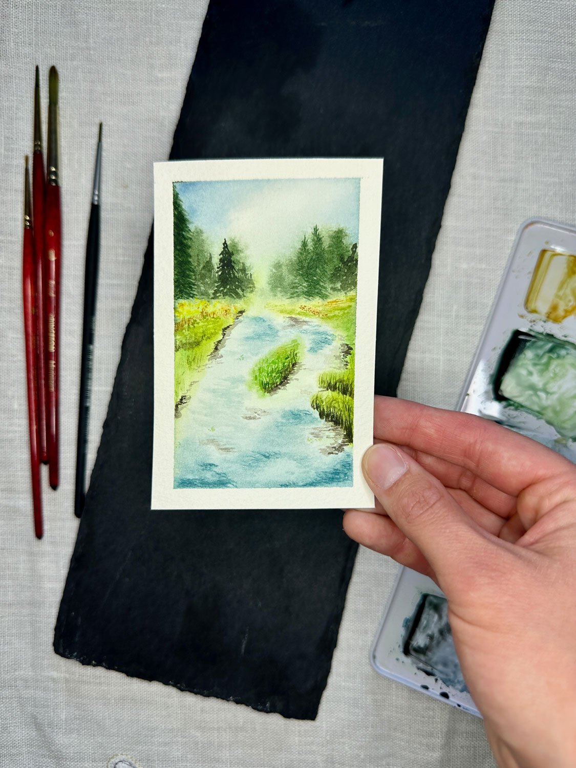 miniature watercolor painting of a creek painted in light blue with pine trees in the background
