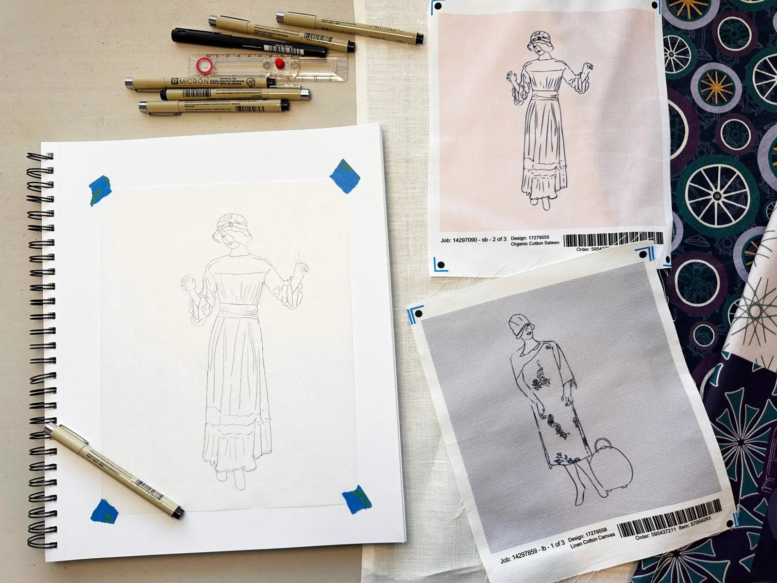 Sketching materials and fashion design illustrations on fabric and paper, including markers, pens, and printed fabric with dress and model sketches.