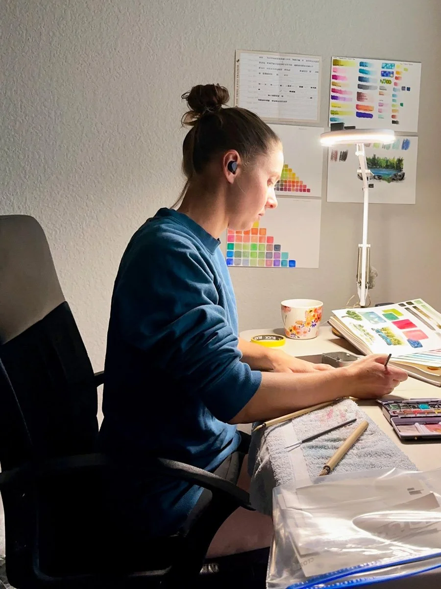A woman sitting at a desk working on watercolor painting, surrounded by color samples, a lamp, and art supplies.