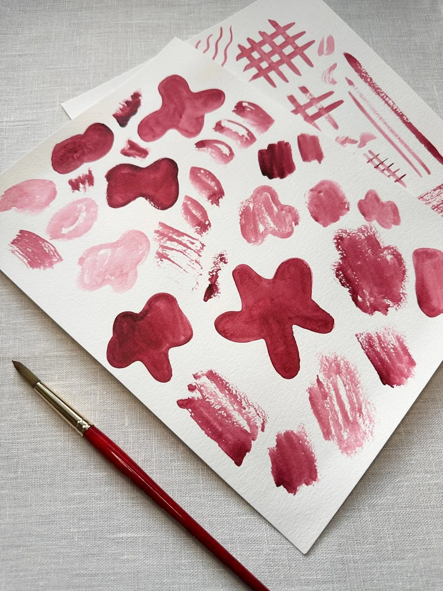 A sheet of white textured paper with various red watercolor paint swatches and shapes, including blobs, strokes, and grid patterns. A red-handled paintbrush is placed nearby on a light fabric surface.