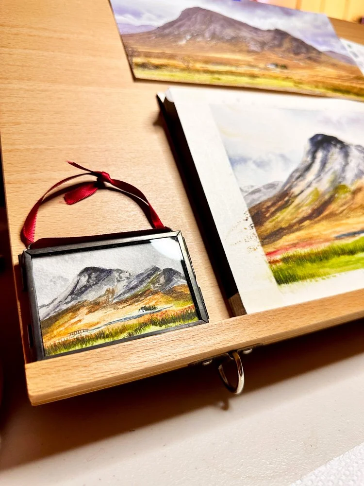 close up of a tiny watercolor ornament in a glass frame next to an in progress larger painting of the same subject.