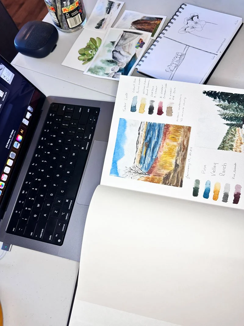 overhead view of a desk with a laptop, watercolor journal, and sketchbook.