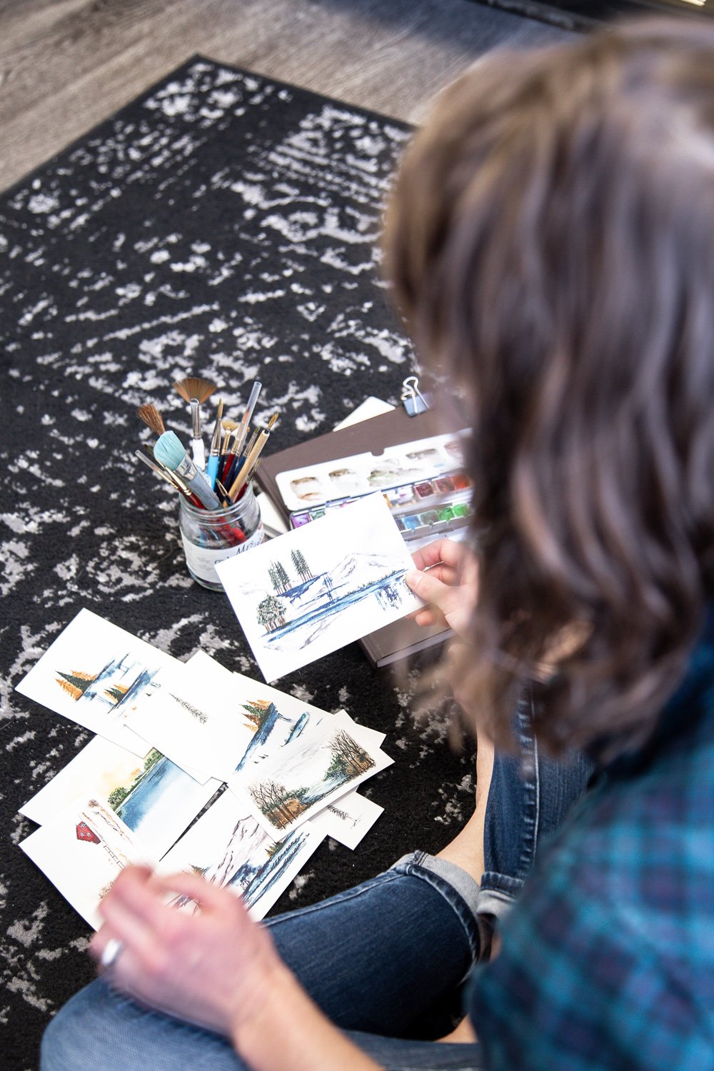 Person with curly hair sitting on a patterned black and white rug, holding a small landscape watercolor painting, surrounded by additional watercolor cards, with brushes and watercolor paints nearby.