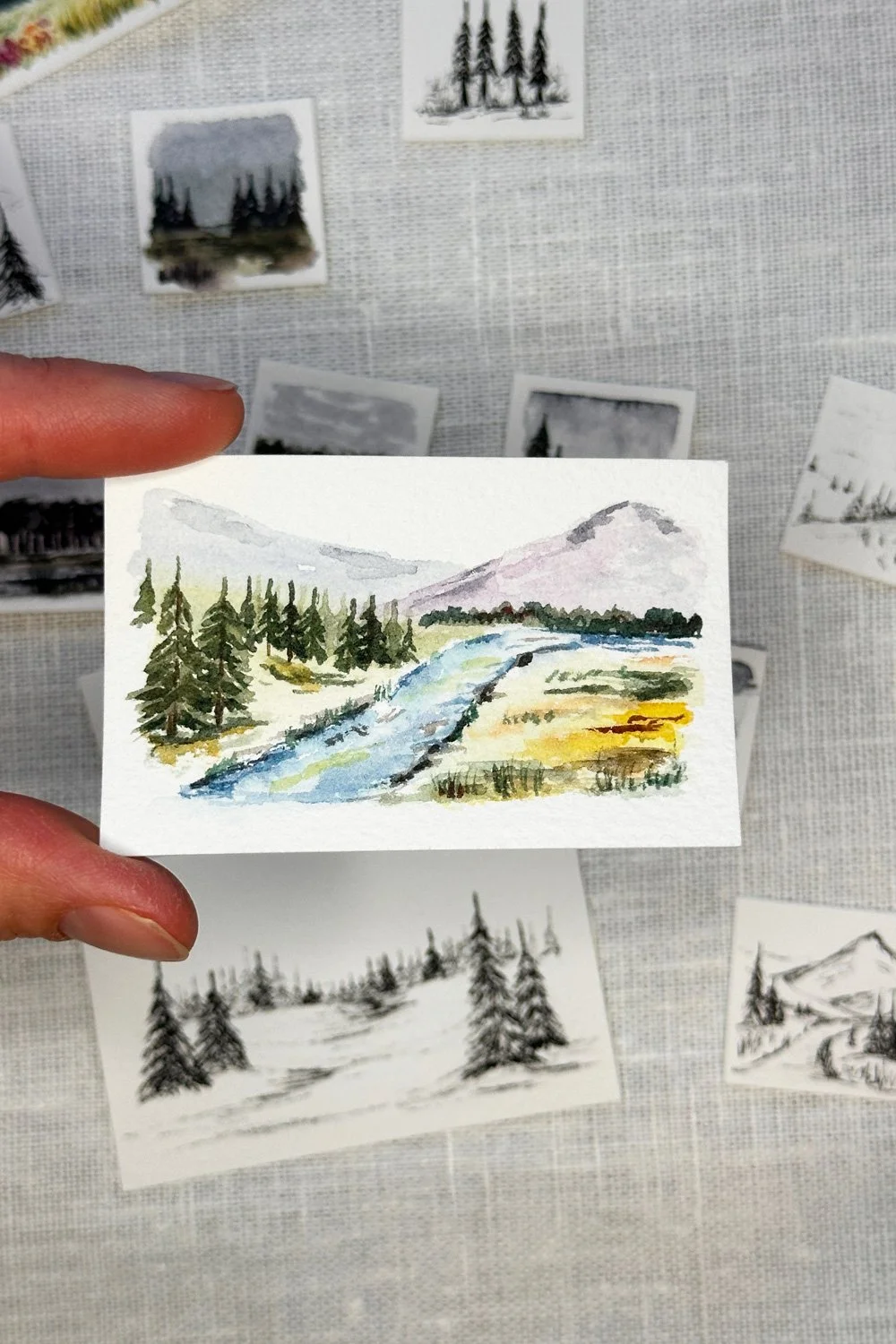 Watercolor painting of a river flowing through a landscape with trees, mountains, and a cloudy sky.