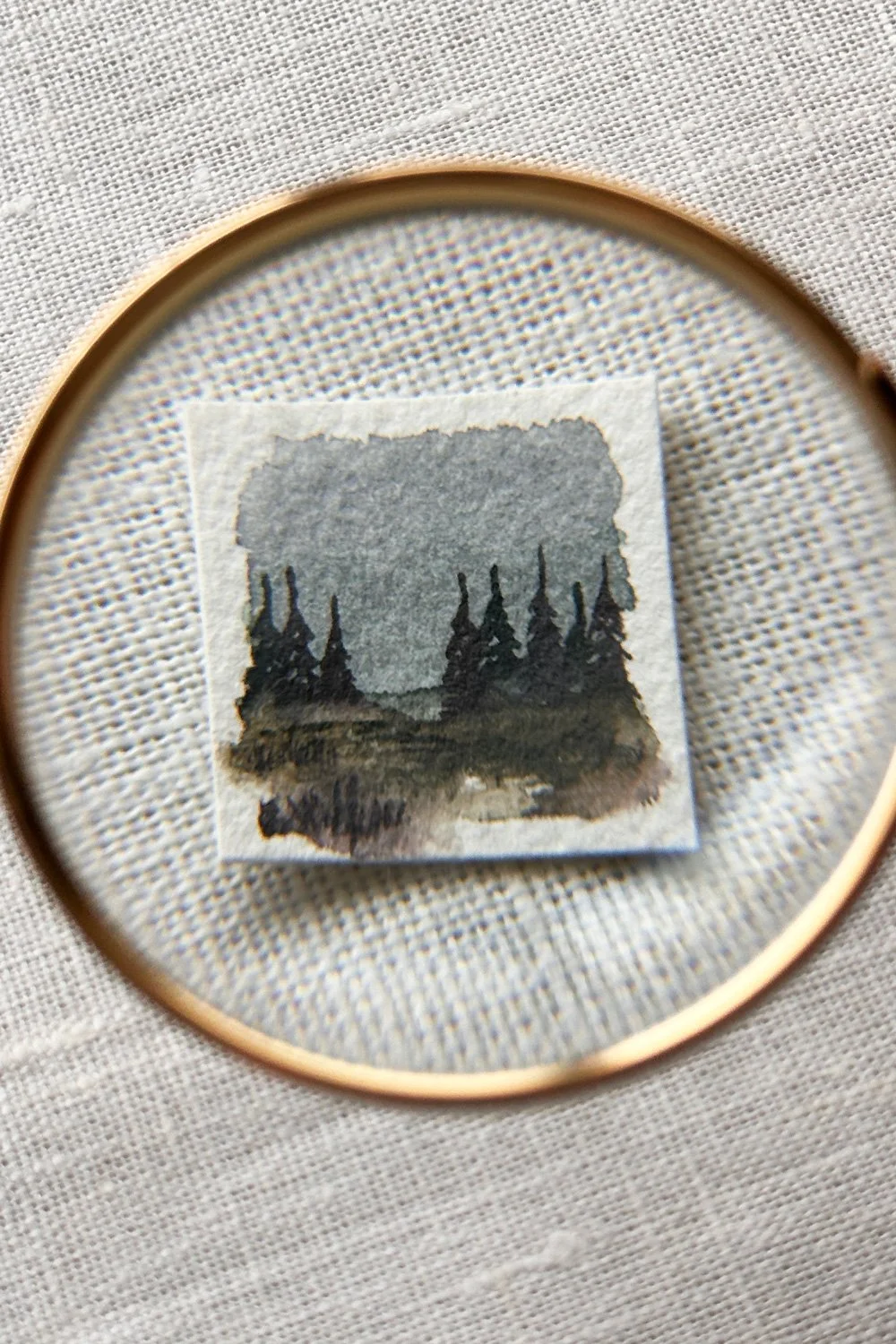Small watercolor painting of a forest landscape with trees and a cloudy sky, viewed through a magnifying glass.