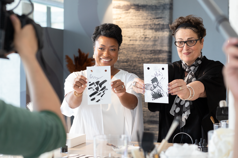 Two women showing black-and-white art prints to a camera inside a modern studio or office space