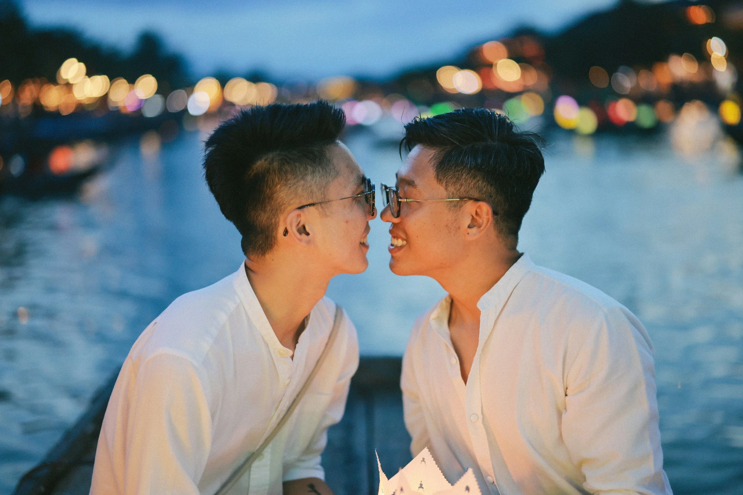 queer couple looking at each other with love, on a dock in front of blue waters