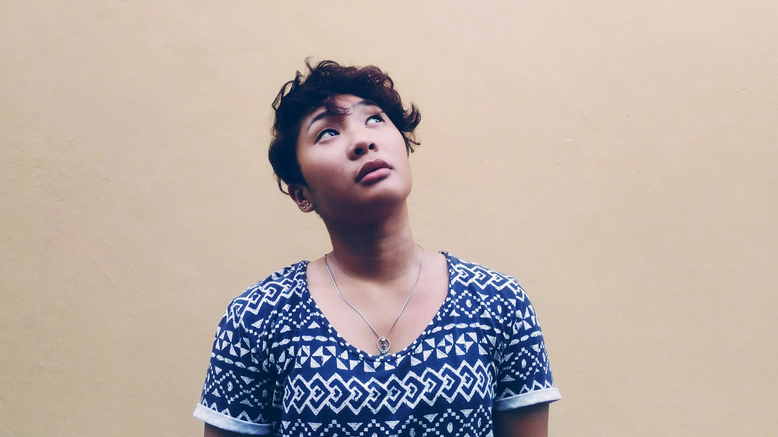 neurodivergent person of color looking upward, wearing blue-patterned shirt & standing in front of beige wall