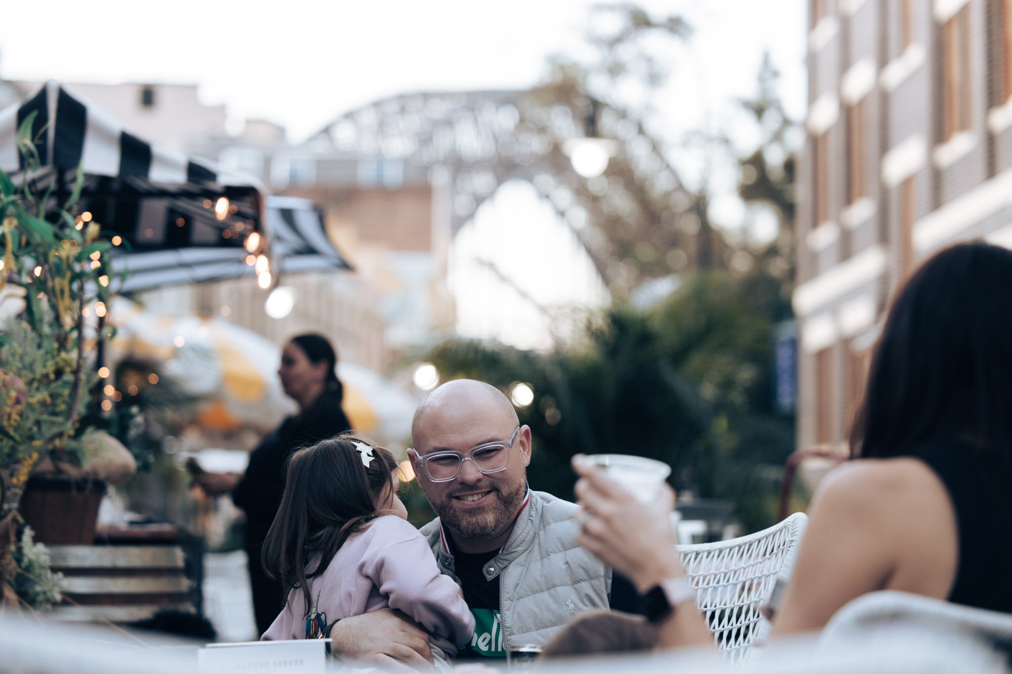 A man with glasses and a beard smiling while holding a young girl, seated at an outdoor cafe with a woman in black sitting nearby, in a city setting with buildings and trees in the background.