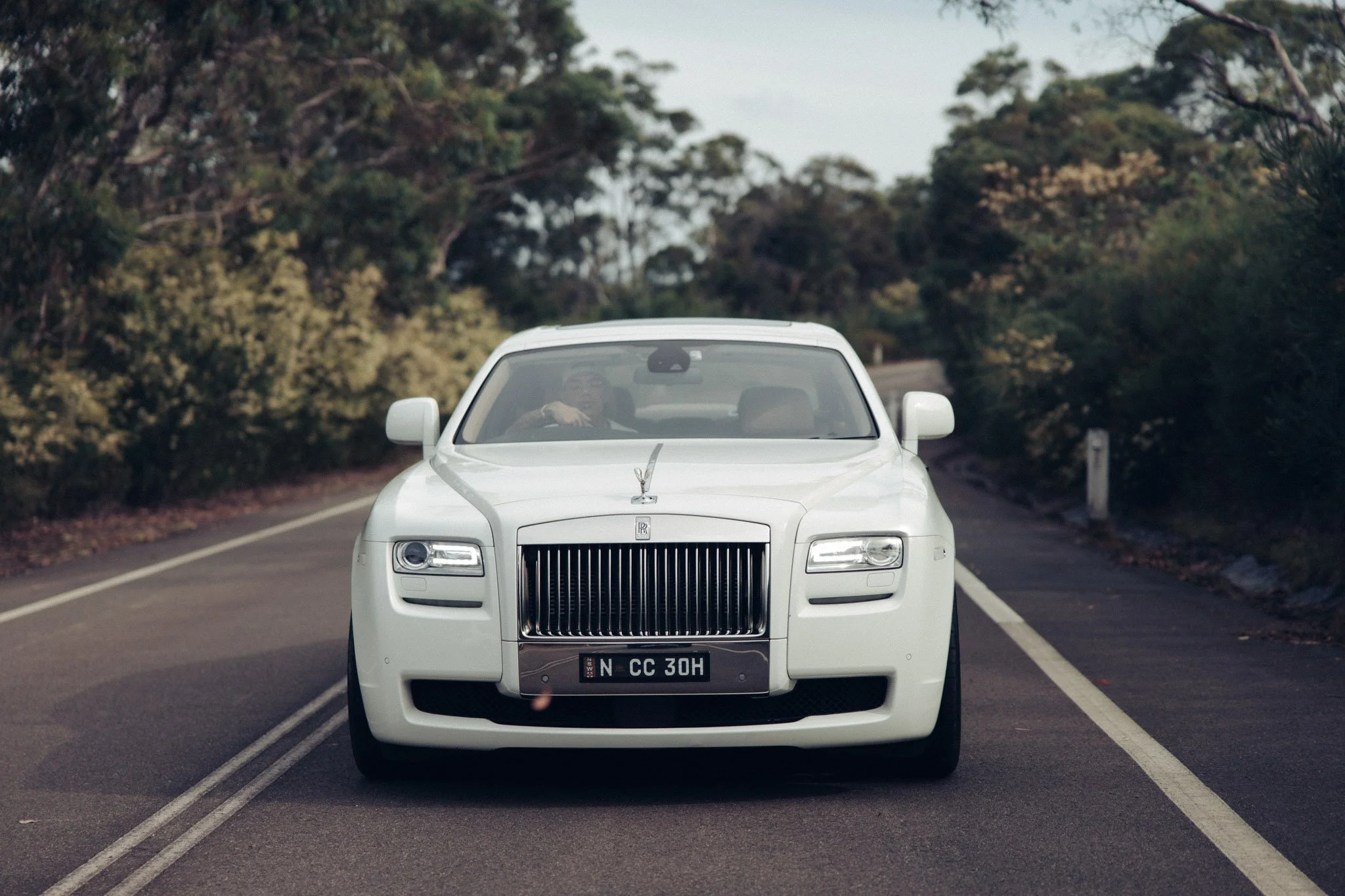 A white Rolls-Royce car driving on a two-lane road surrounded by trees.
