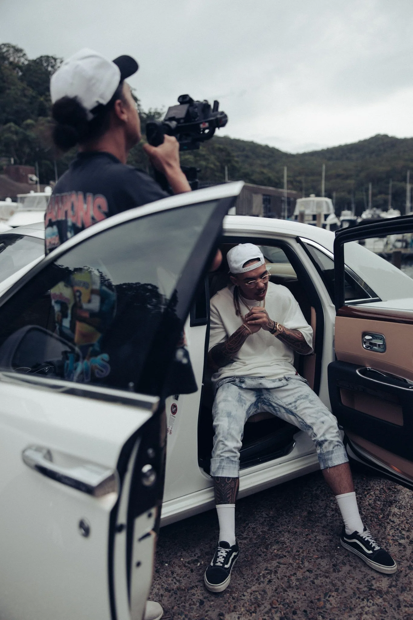 A man with tattoos sits in a luxury car with its door open, wearing a white cap backwards, white t-shirt, and ripped jeans, while a cameraman filming him stands outside the car.