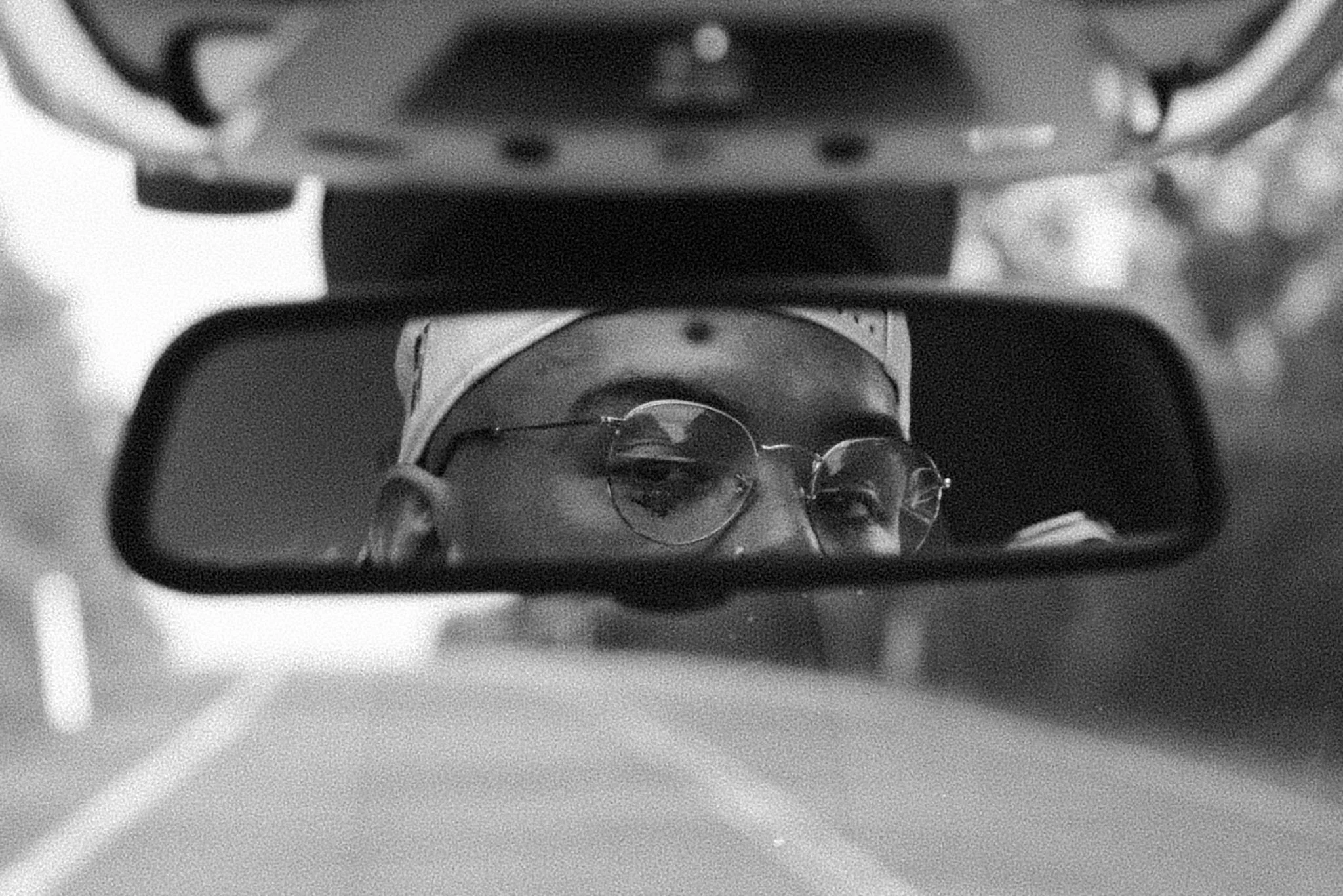 A person with glasses and a bandana looking into a rearview mirror, with part of a vehicle's dashboard visible.