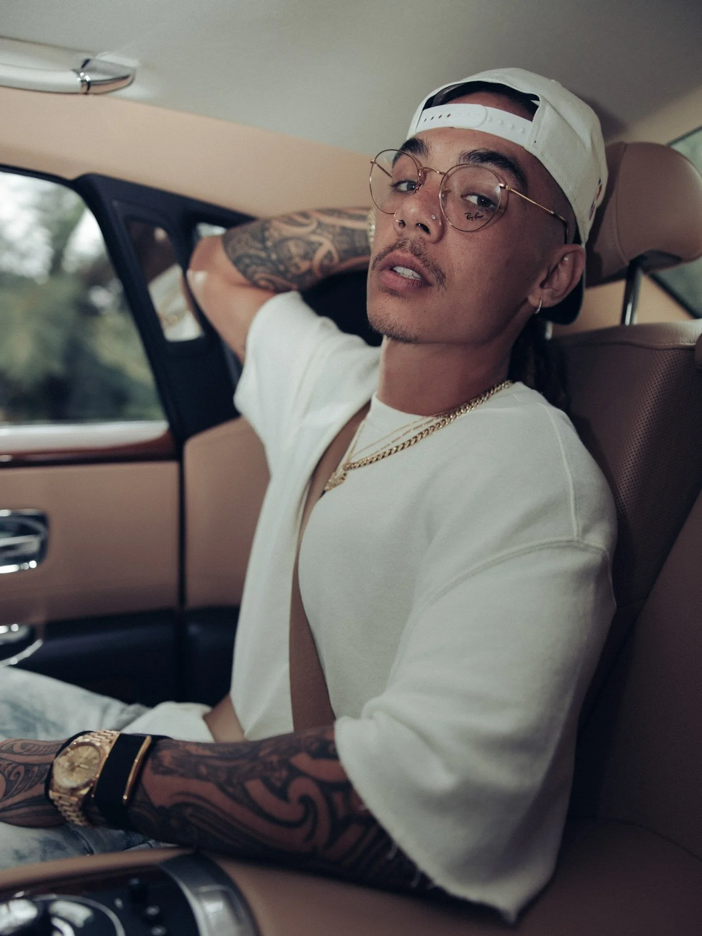 A young man with tattoos, glasses, and a nose piercing sitting in the backseat of a car, wearing a white cap backwards, a white t-shirt, a gold chain, and a watch.