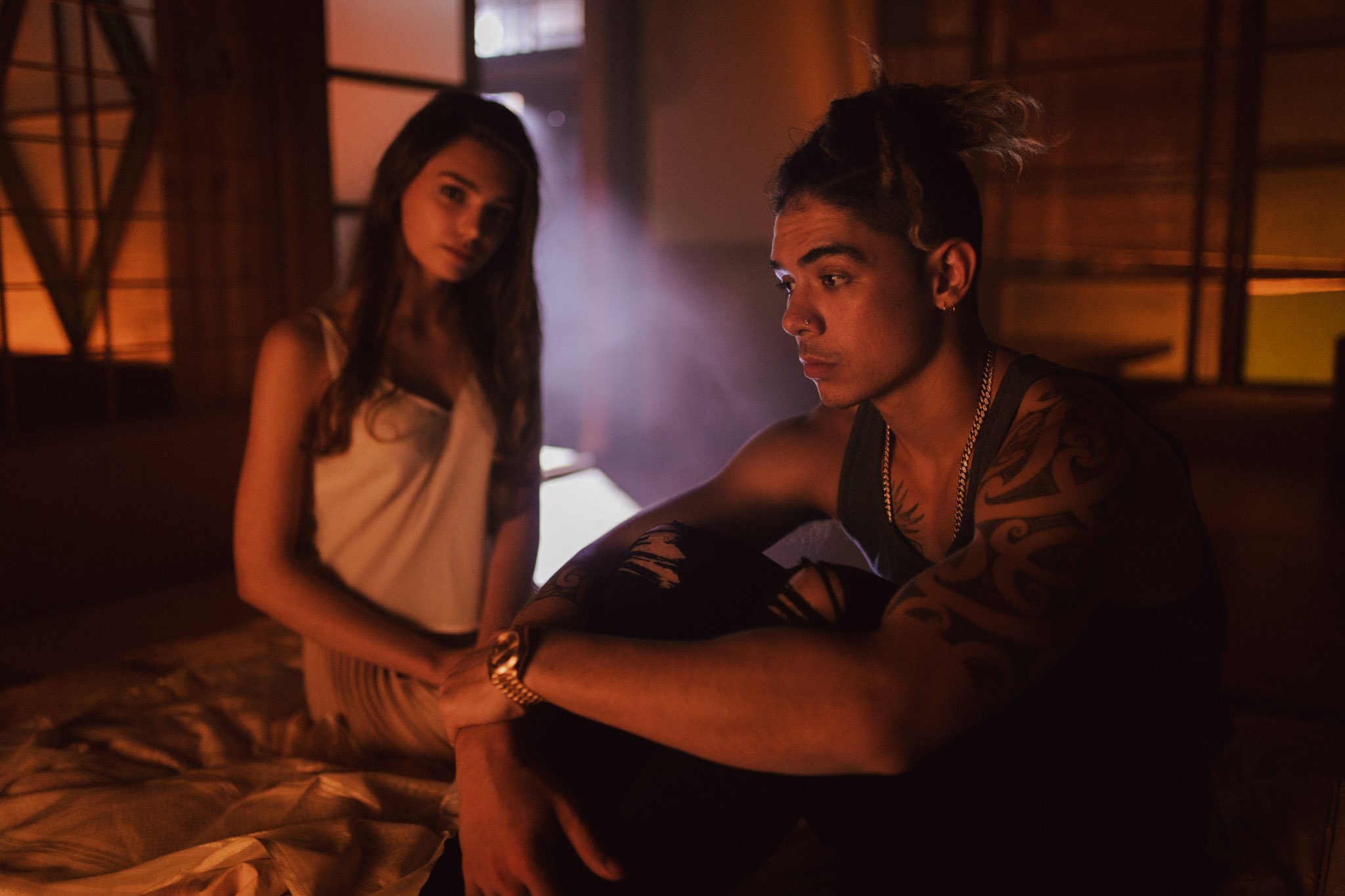 A young woman and a young man with tattoos sitting in a dimly lit room with warm orange lighting, wooden walls, and a large window, creating a moody atmosphere.