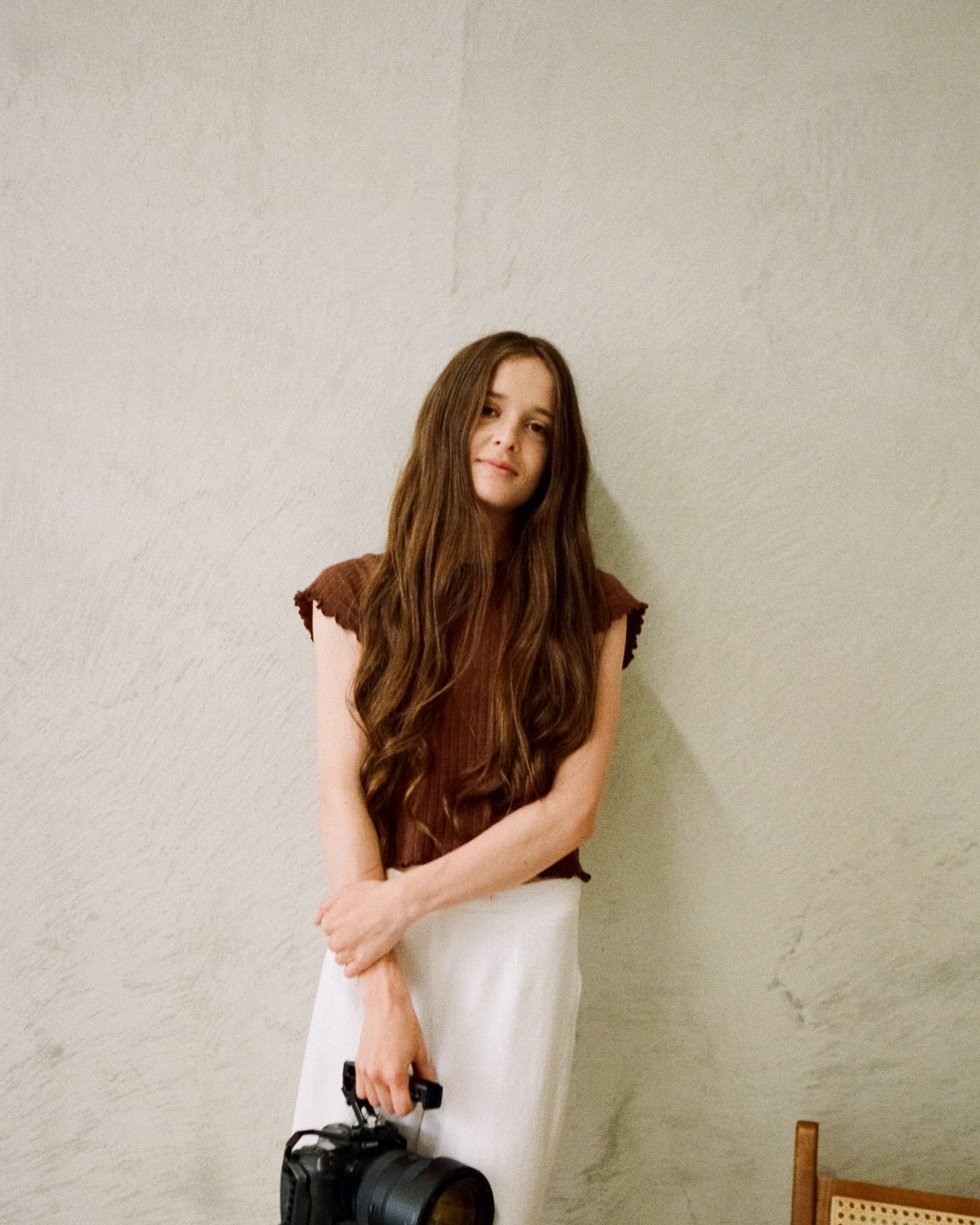A young woman with long brown hair wearing a rust-colored top and white skirt, holding a camera, standing against a textured beige wall.