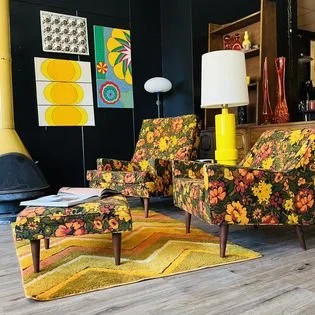 2 mid century modern floral armchairs, mid century modern yellow table lamp, black and red  swung vases, abstract wall art, and a yellow rug.