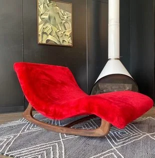 Adrian Pearsall Double wave rocking chaise lounger, situated on a geometric-patterned rug in a room with dark walls, a white mid century modern preway fireplace, and a modern abstract art piece on the wall.