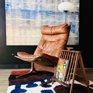 Brown leather Westnova Siesta lounge chair with wooden frame next to an Author Umanoff  magazine rack / table holding magazines,  in a studio space with a large original painting by James Adley and a Laurel mushroom floor lamp, on a  Marimekko  rug.