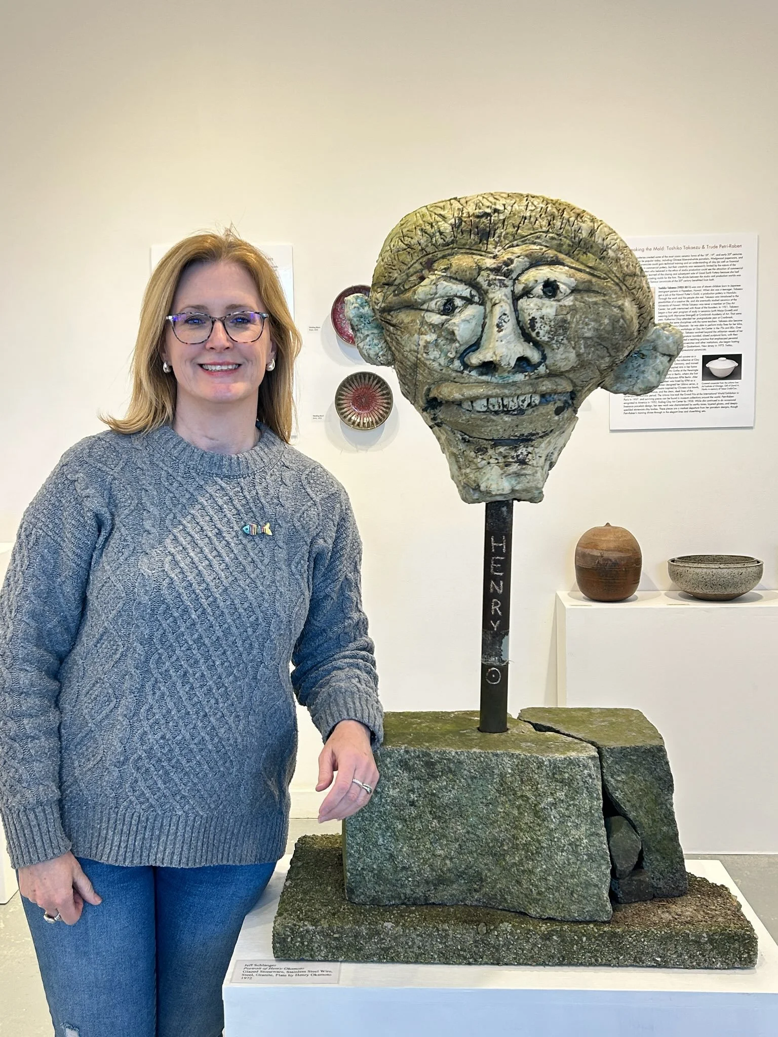 A woman wearing a grey sweater stands next to a clay sculpture of a head in a gallery.
