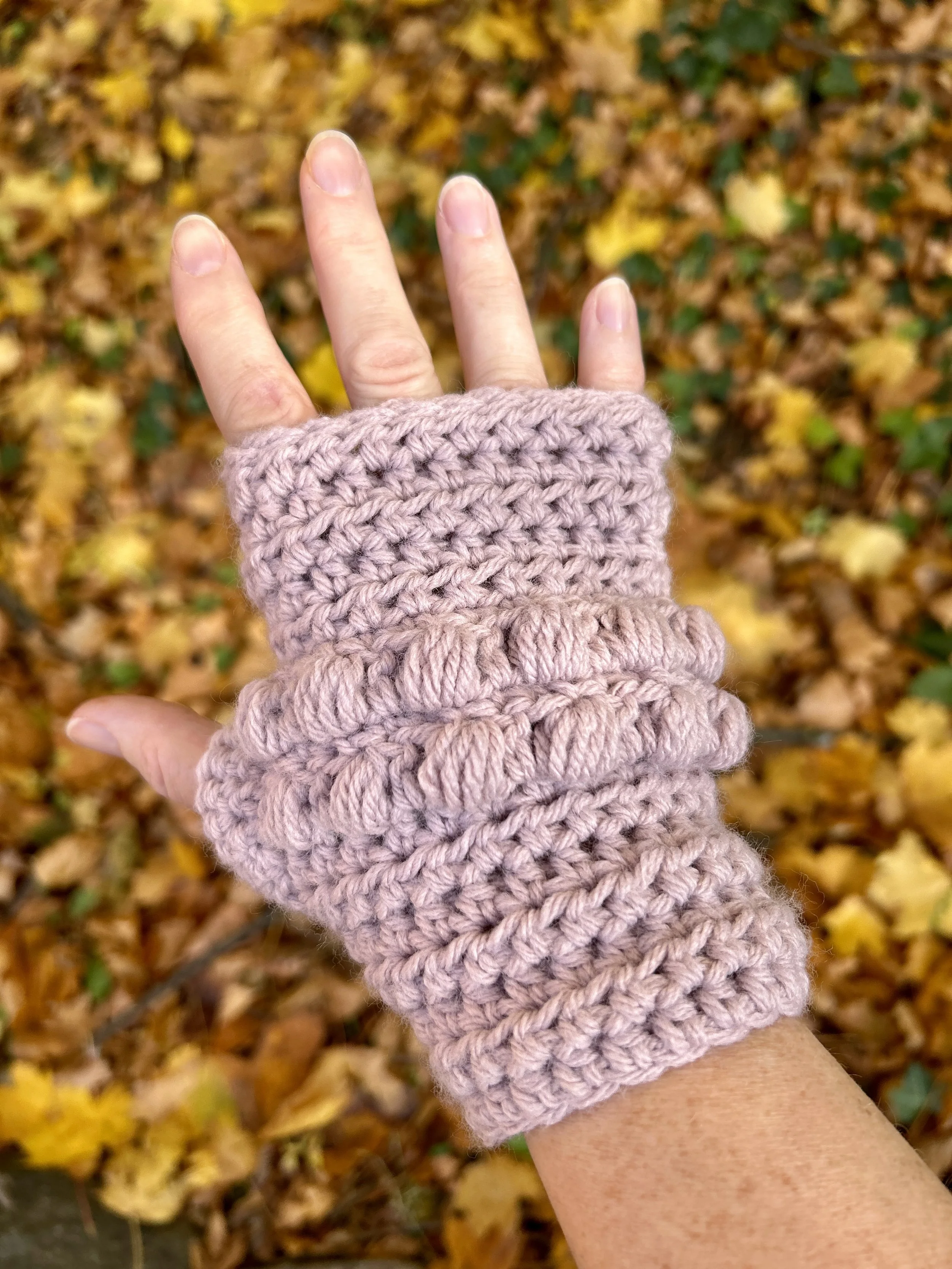 A person wearing pink crochet fingerless gloves with a background of yellow autumn leaves.
