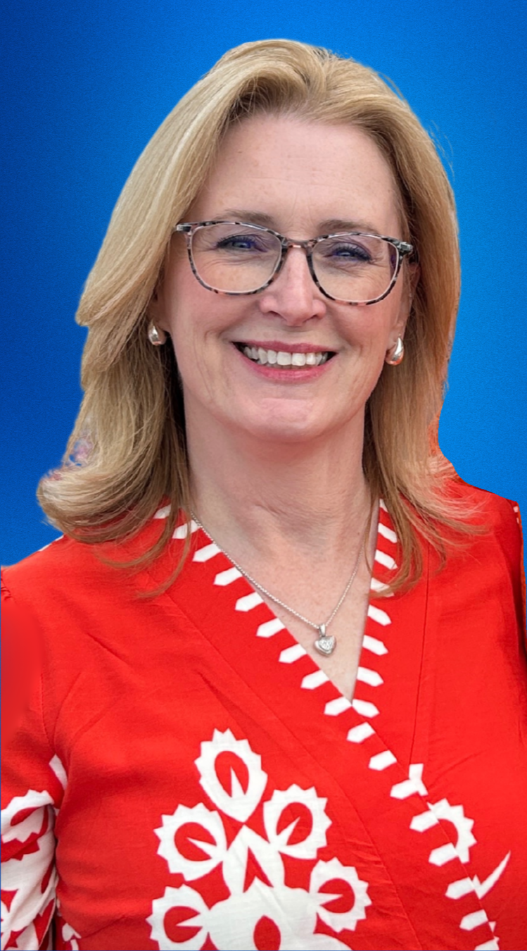 A smiling woman with blonde hair wearing glasses, a red and white patterned dress, a necklace, and earrings, standing against a blue background.