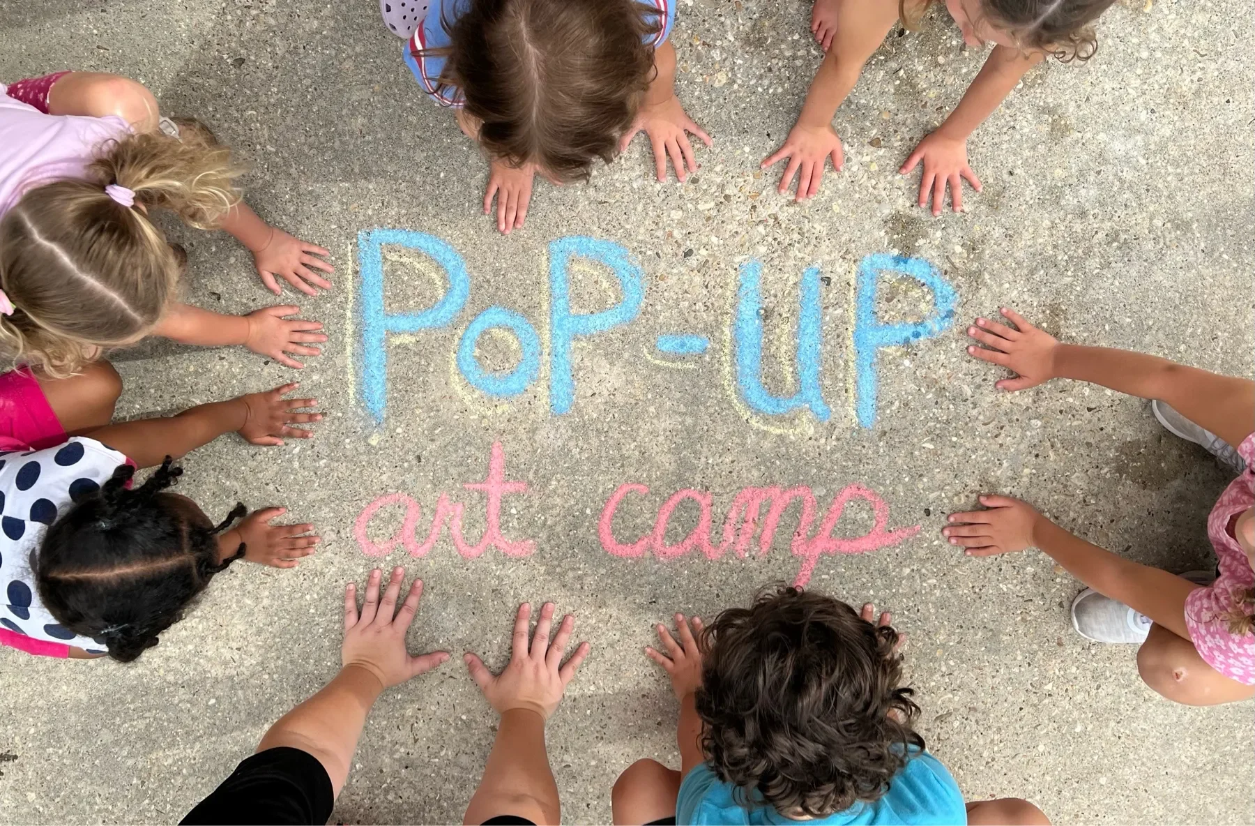 Students and their art teacher are bending down and placing their hands around a chalk-drawn image that says, "Pop-Up Art Camp." This is the cover image for the SEO blog that describes art camp and invites parents to register their children.
