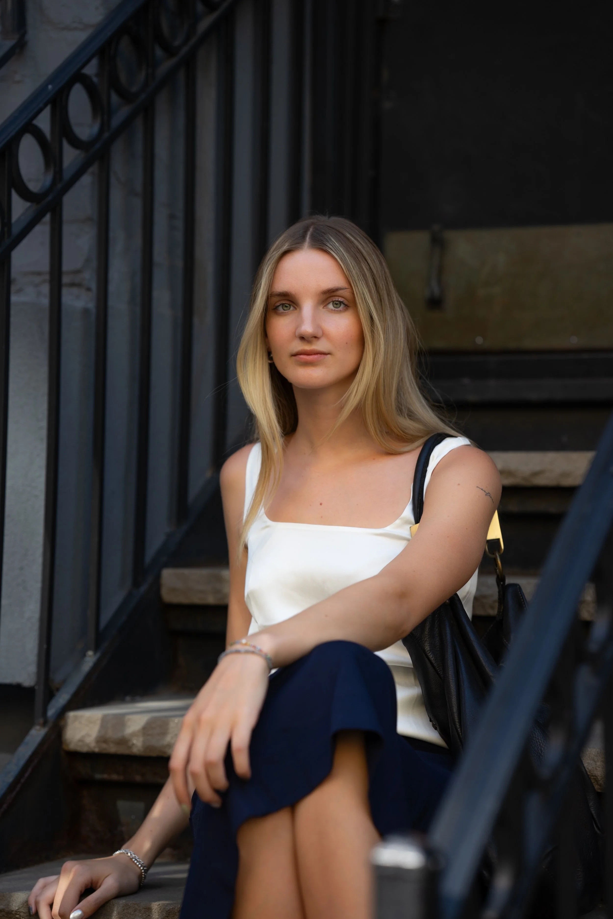 A young woman with blonde hair sitting on outdoor stairs, wearing a white top and dark pants, carrying a black bag over her shoulder.