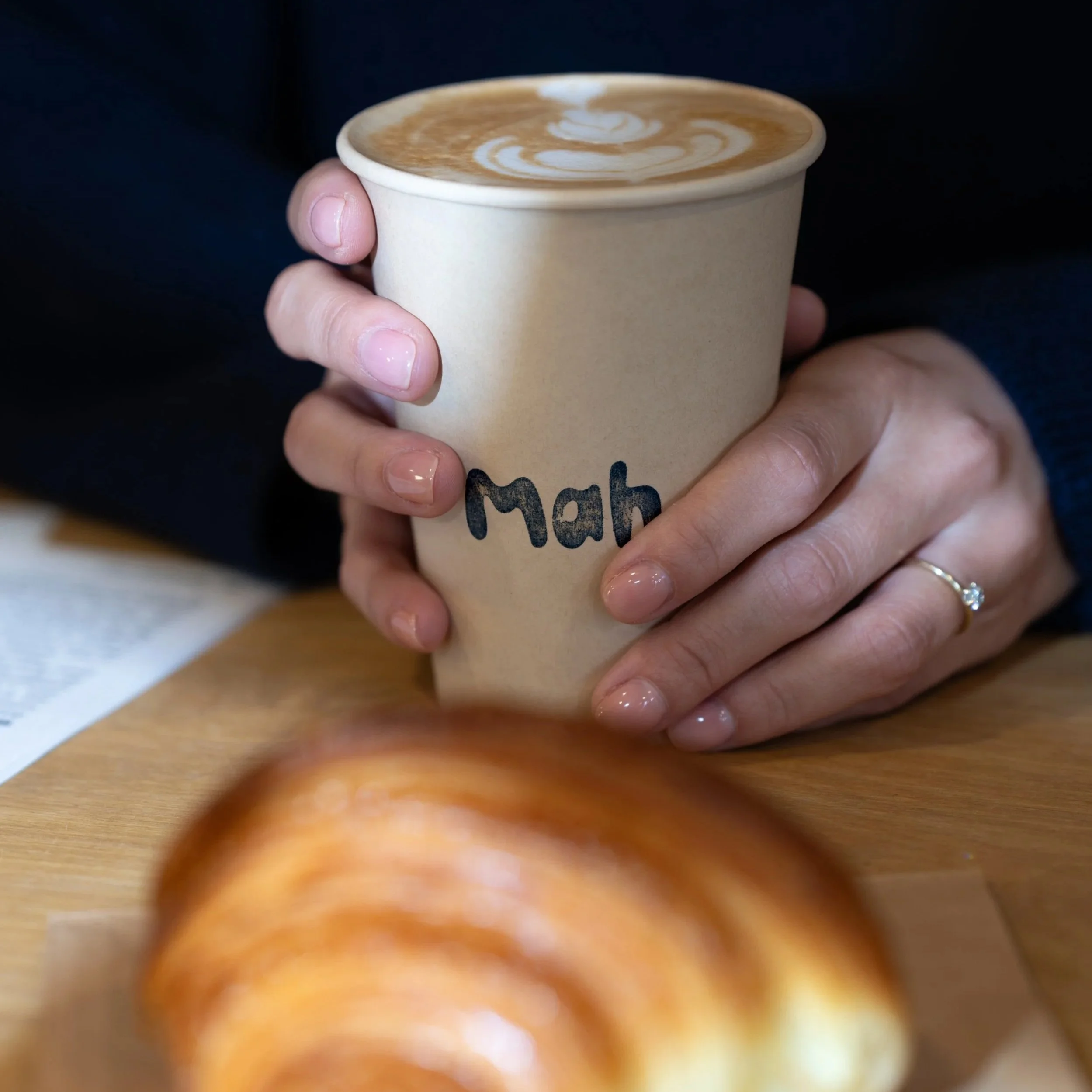 A person holding a beige paper cup with 'Mab' written on it, containing a latte with a swirl foam design, seated at a wooden table with a pastry in the foreground.