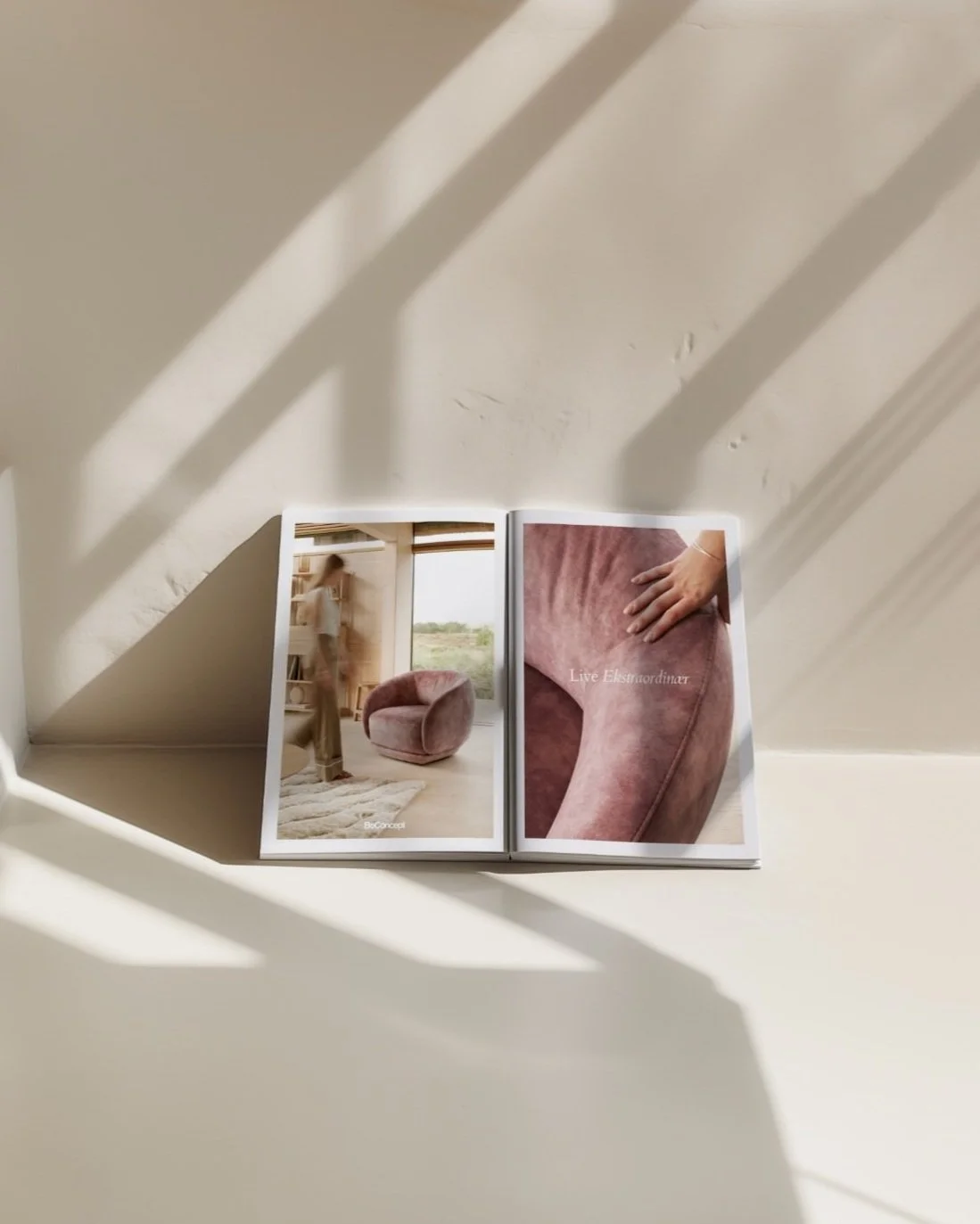 An open magazine displaying interior design photos of a pink velvet chair and a hand touching the chair's armrest, placed on a white surface with sunlight and shadow patterns.