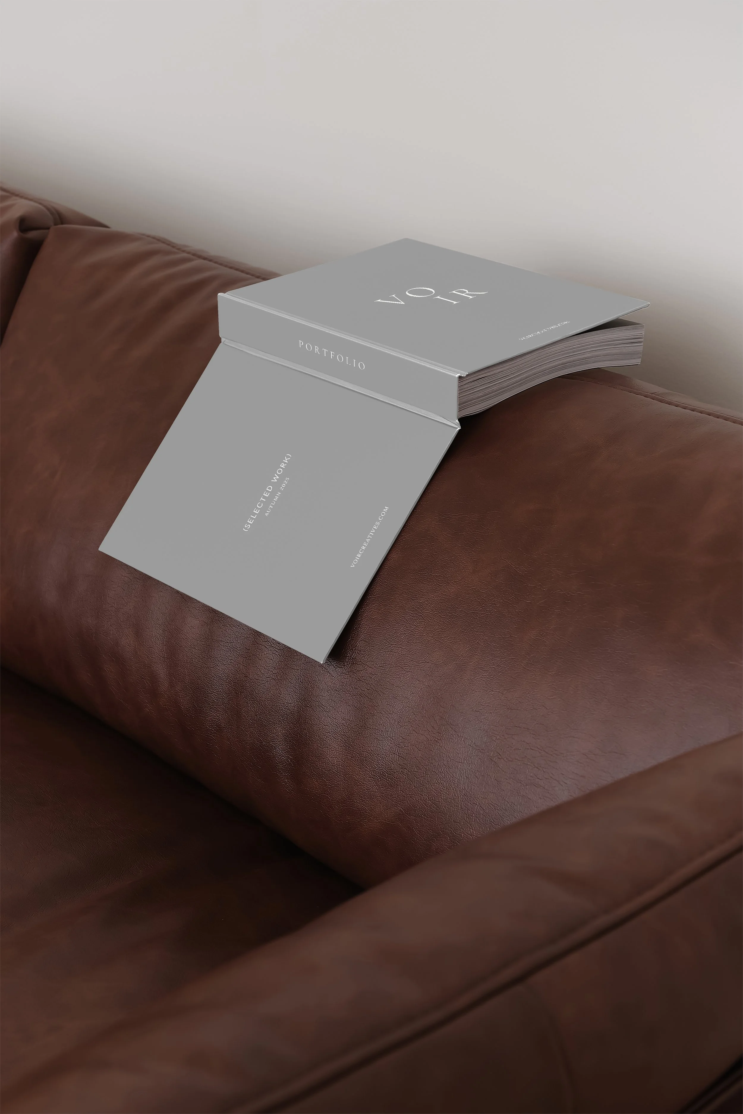 A brown leather couch with a gray portfolio and a matching gray folder placed on it. The folder displays the text 'Selected Work' and the portfolio has the words 'VOR Portfolio' on its cover.