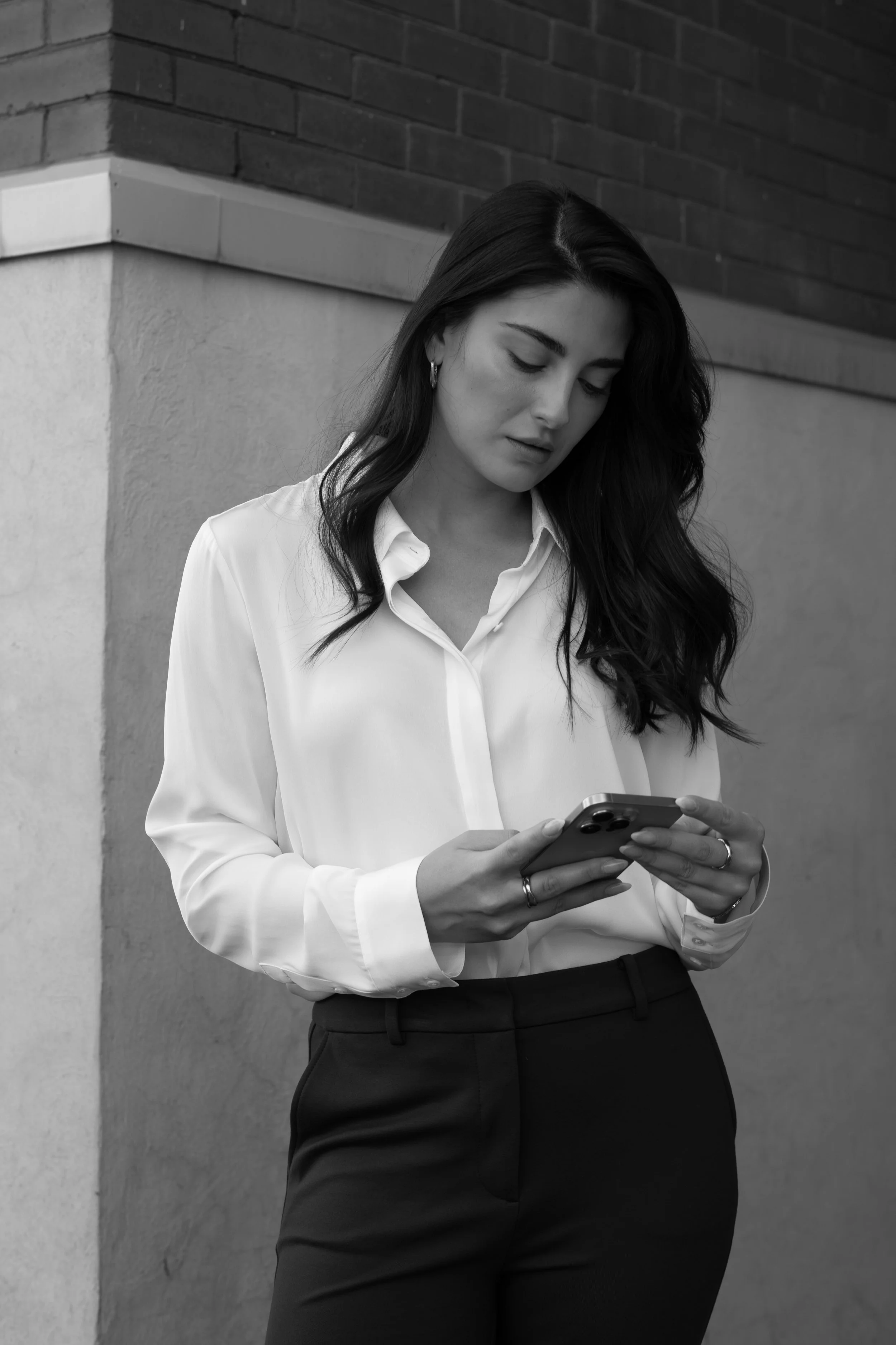 A woman in a white blouse and black pants standing against an outside concrete wall, looking at her phone.