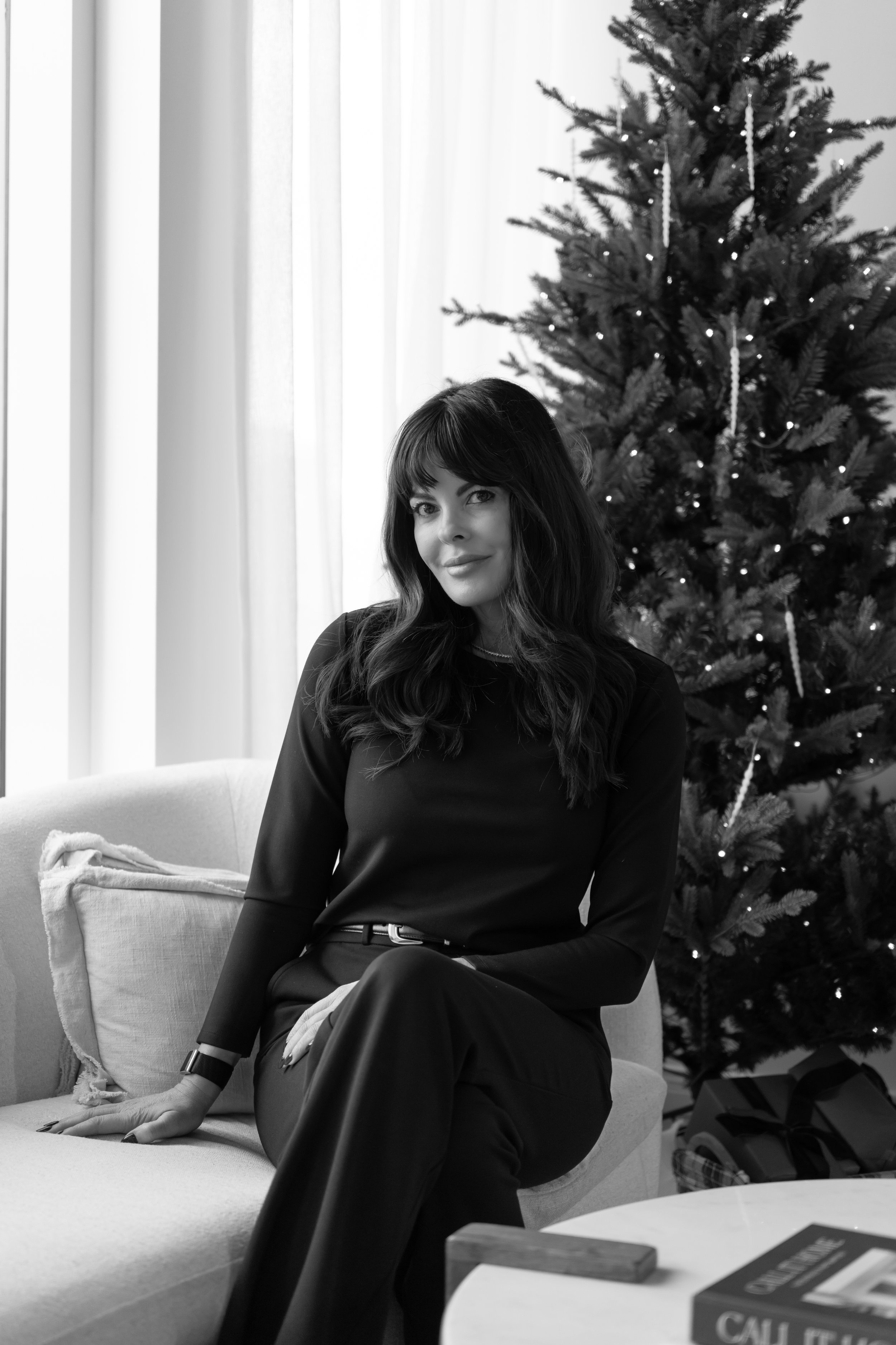 A woman with dark wavy hair sitting on a sofa near a decorated Christmas tree.