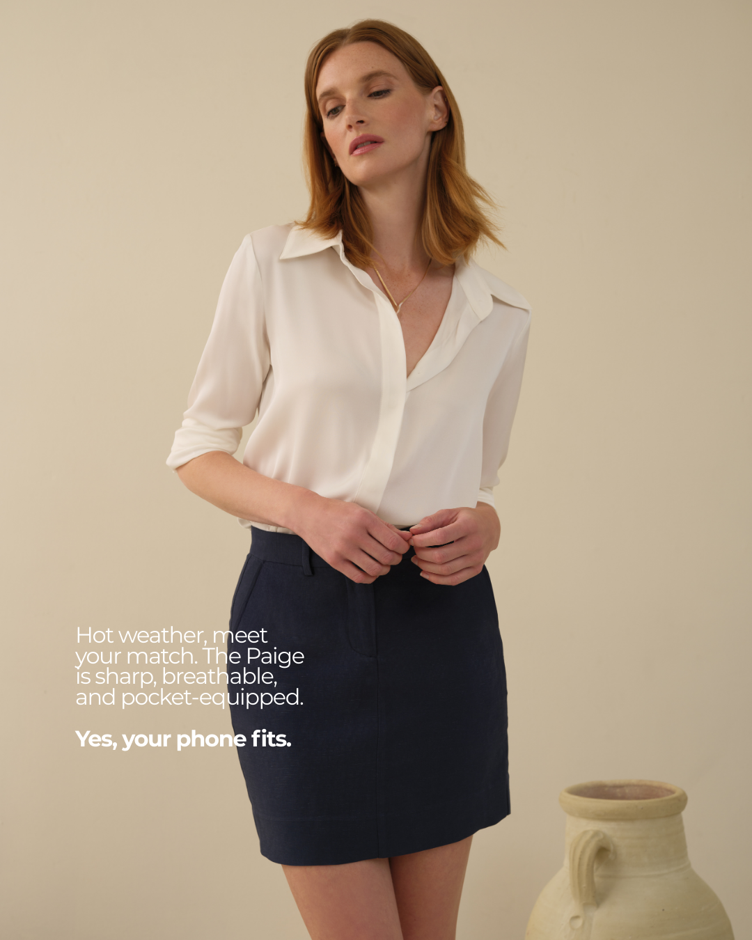 A woman with red hair wears a white blouse and a navy skirt, standing next to a pottery vase. The image promotes a breathable, pocket-equipped shirt that fits your phone.