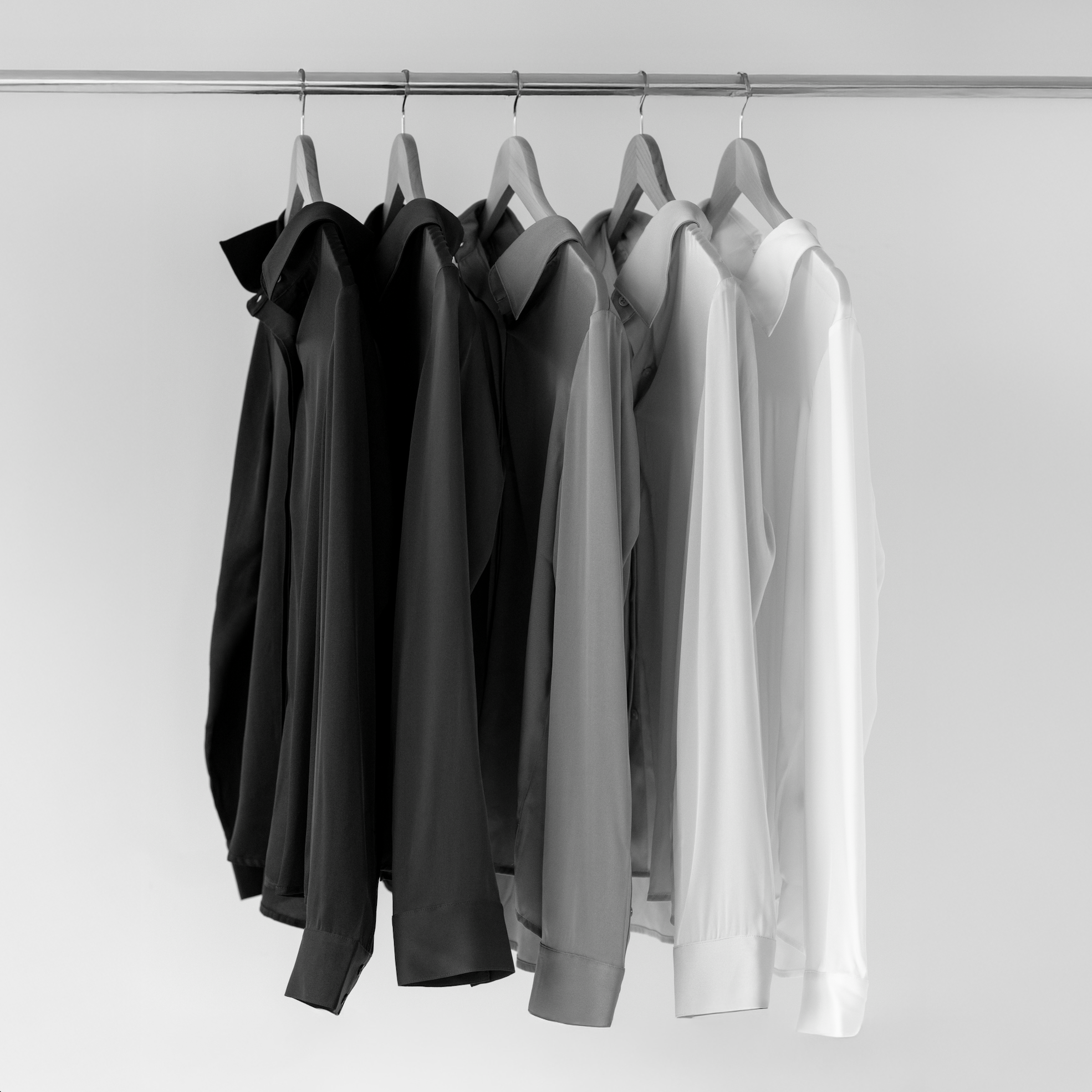 Six hanging shirts arranged from black to white on wooden hangers against a plain wall.