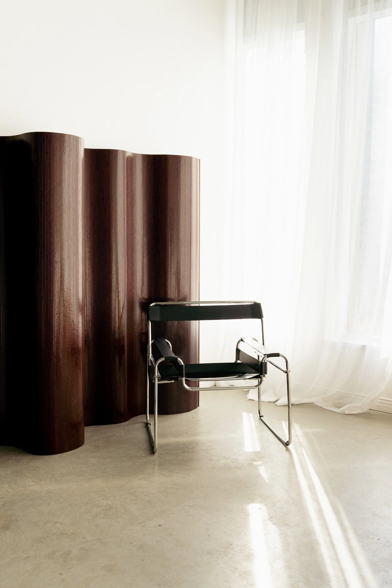 Chrome and black chair beside a sculptural wooden screen in natural window light at Studio by VOIR.