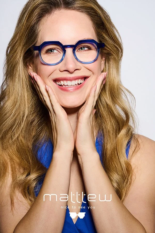A woman with long wavy blonde hair, wearing blue glasses and a blue top, smiling with hands on her cheeks.
