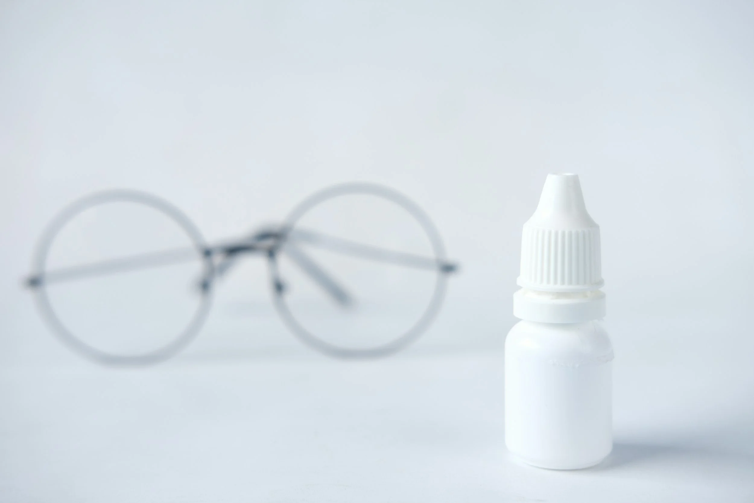 A pair of blurry eyeglasses and a white eye drop bottle on a white surface against a plain white background.