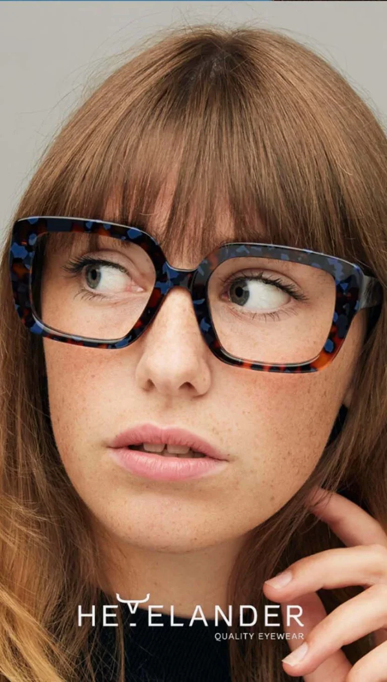 Close-up of a woman with red hair wearing tortoiseshell eyeglasses, looking to her right with a thoughtful expression. Text overlay reads "HEYEANDER QUALITY EYEWEAR."