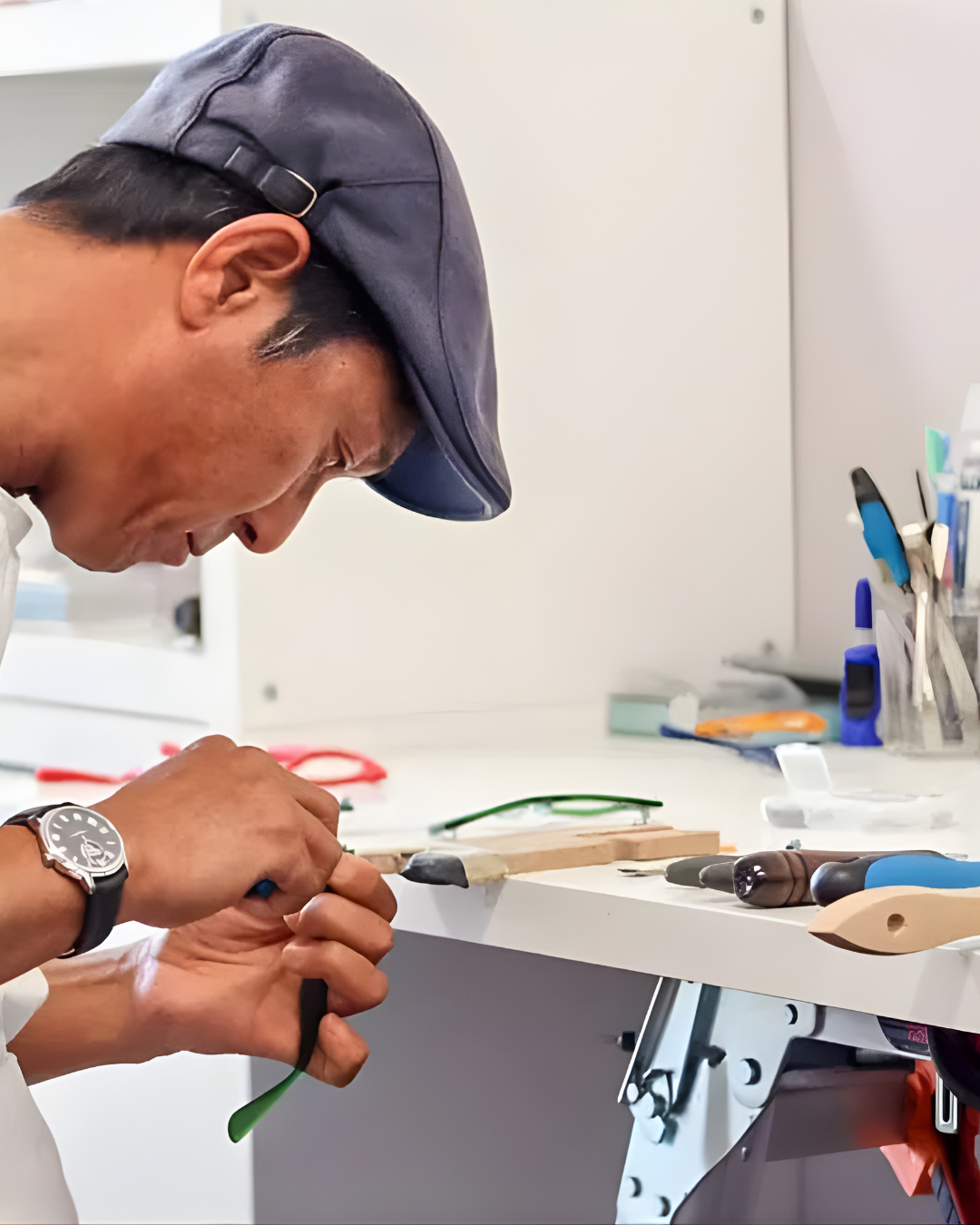 A man working at a cluttered workstation with various tools on a white table, wearing a dark gray cap and a wristwatch.