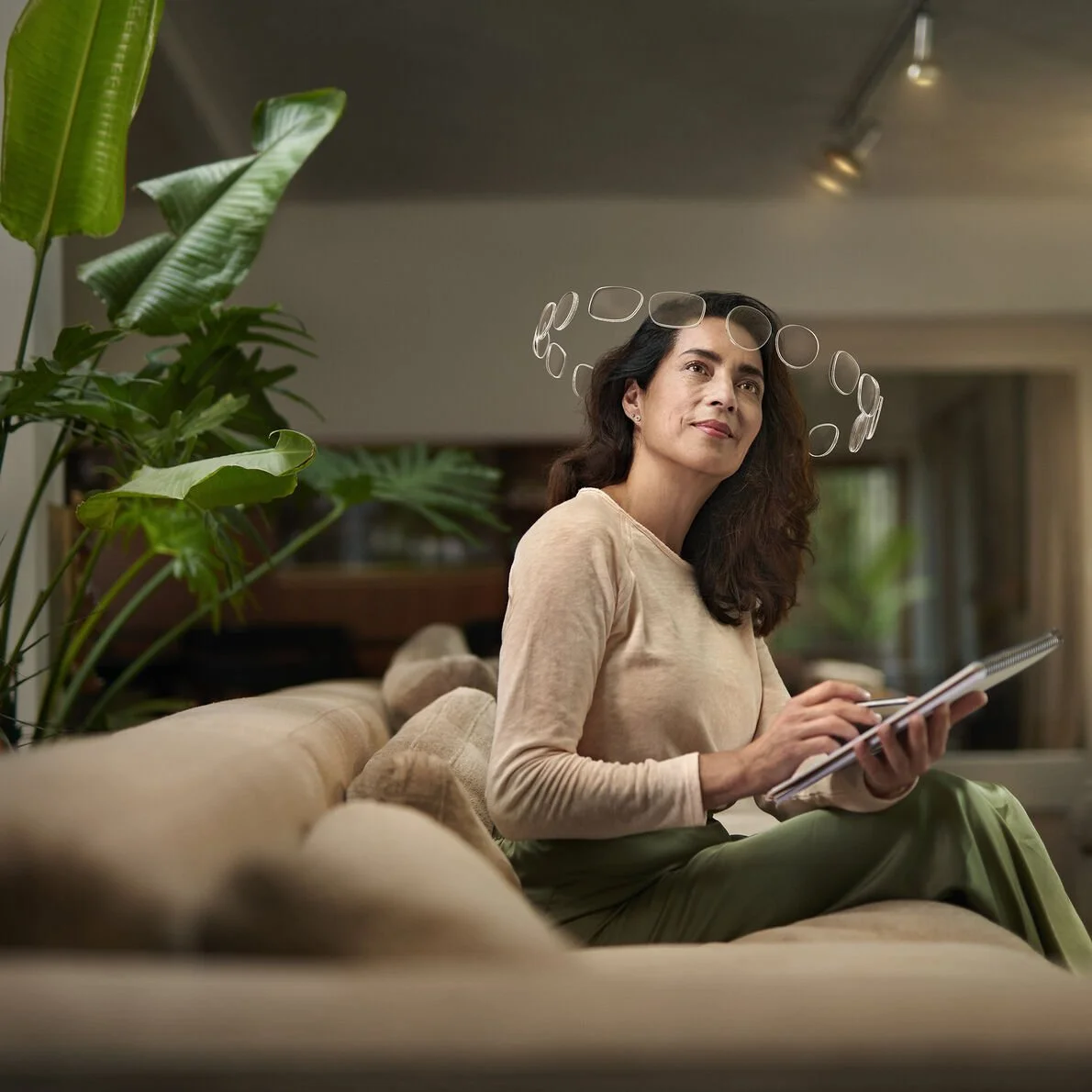 Woman sitting on a beige sofa with a notebook, surrounded by plants in a cozy living room.